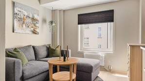 A modern living room with a grey sofa, a round wooden table, and a large window with a view of buildings. - Cozy Studio in Bergen Center (Bergen)