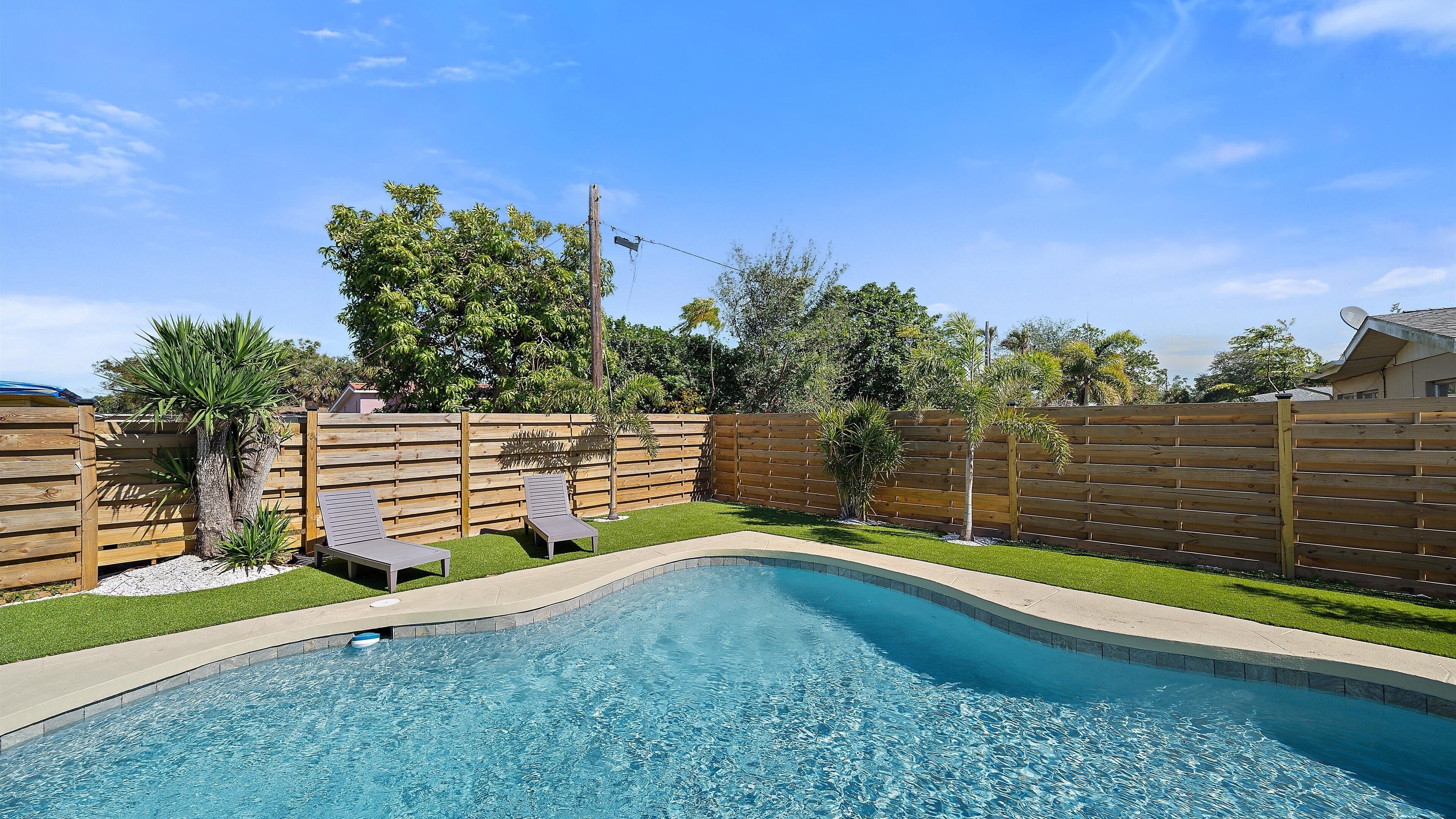 A backyard with a swimming pool, two lounge chairs, a wooden fence, and a variety of trees.