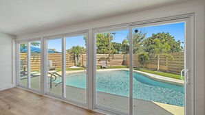 A spacious room with a large sliding glass door leading to a backyard with a swimming pool, wooden fence, and outdoor furniture. - Coastal Cottage 2 Bedroom Home (Sarasota)