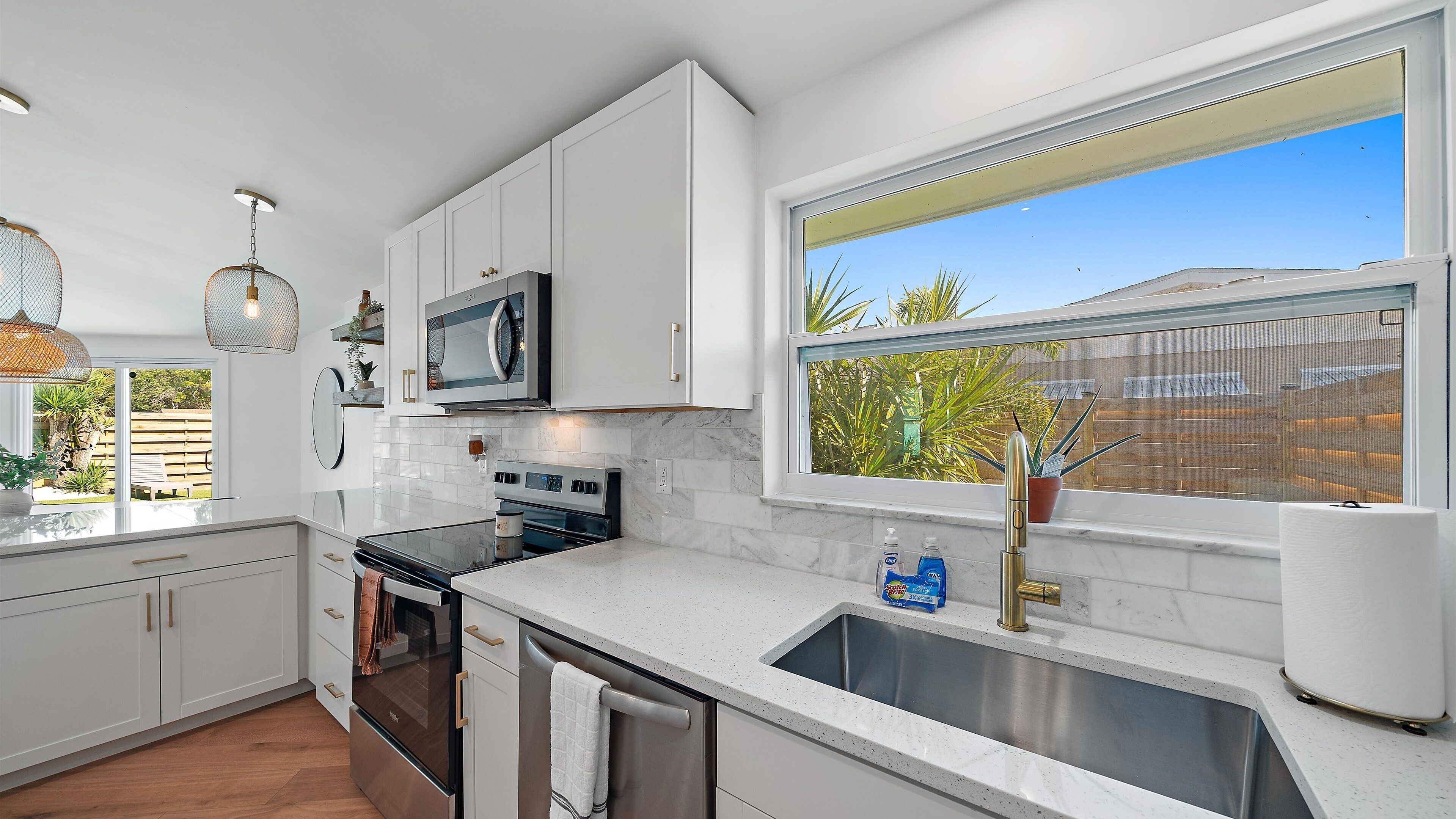 A modern kitchen with white cabinetry, stainless steel appliances, a large window with a view of the outside, and a countertop with a sink and a bottle of soap.
