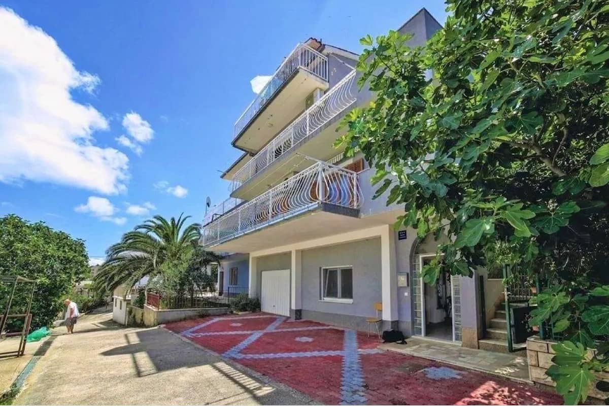 A multi-story residential building with balconies, a red patterned driveway, and lush greenery.
