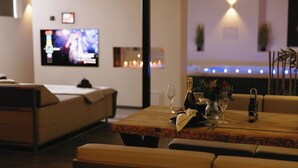 A modern living room with a sofa, a wooden coffee table with a vase, wine glasses, and a bottle. A flat-screen TV is mounted on the wall, and there are decorative lights on the ceiling. - Waal Wellness Villa Maas and Waal Wellness & Suites (Dreumel)