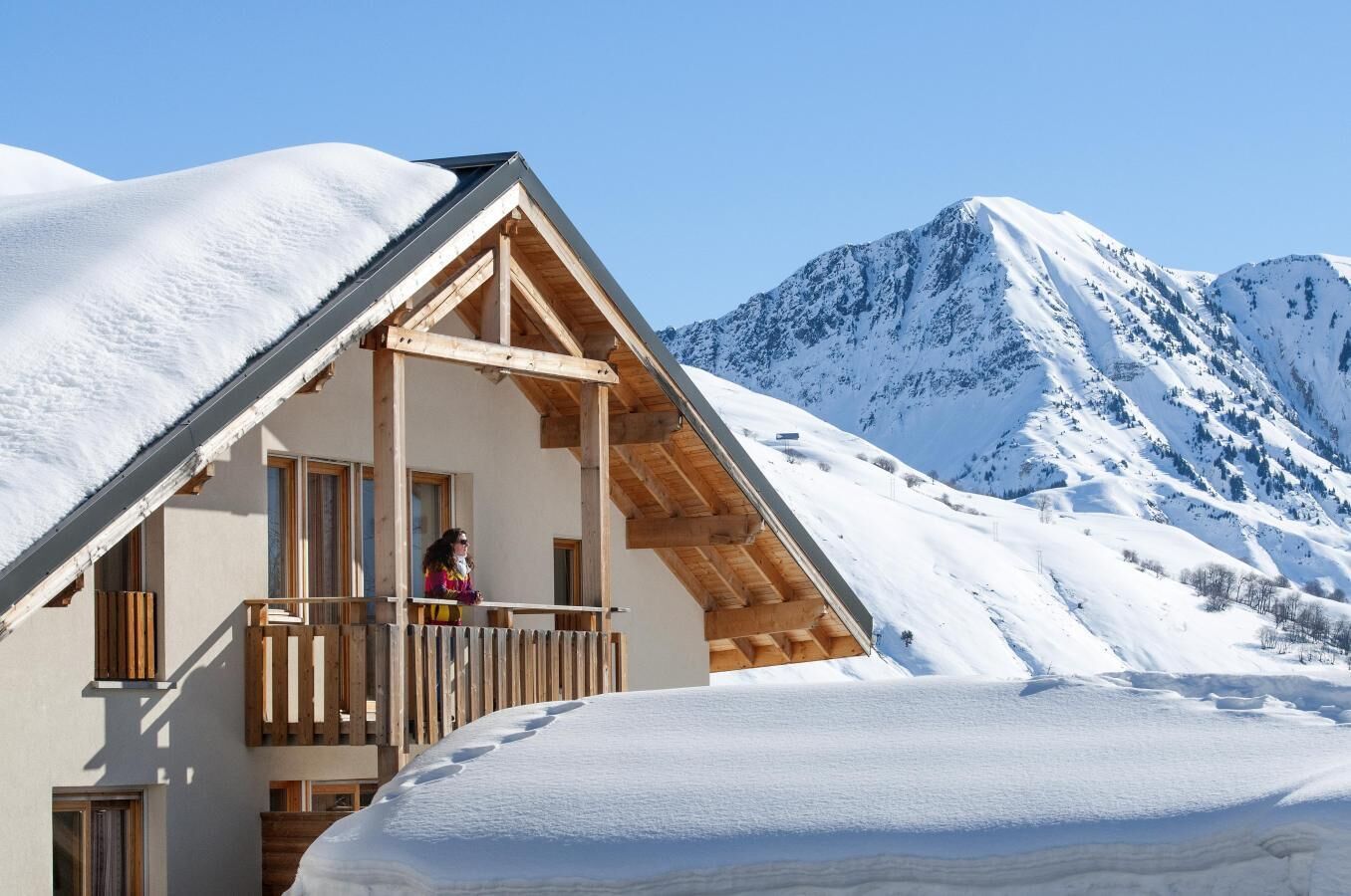 A chalet with a wooden balcony, a mountain backdrop, and snow-covered surroundings.