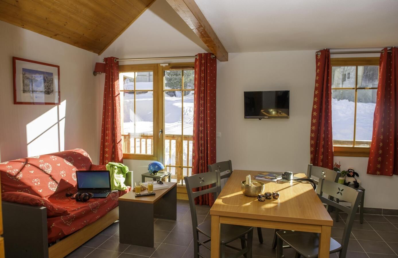 Ein gemütliches Zimmer mit Holzbalkendecke, einem Sofa mit Laptop, einem Esstisch mit Stühlen und einem Fernseher an der Wand.