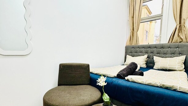 A modern bedroom with a large bed, two ottomans, a potted plant, and a view of buildings through the window. - Cozy Rooms Superb Location near Augarten (Vienna)