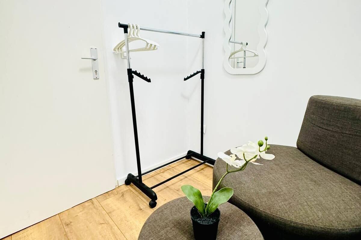 Una habitación con piso de madera, un sofá gris, un otomanillo redondo con una orquídea en maceta, un tendedero metálico y un espejo fijado a la pared.