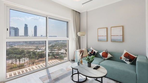 A modern living room with a large window, a green sofa, a coffee table, and two framed artworks on the wall. - Livbnb-Modern Living 1B Apartment in JVC (Dubai)