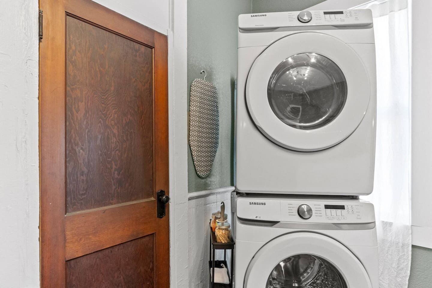 A laundry area with two Samsung washing machines stacked, a wooden door, and a decorative item on the wall.