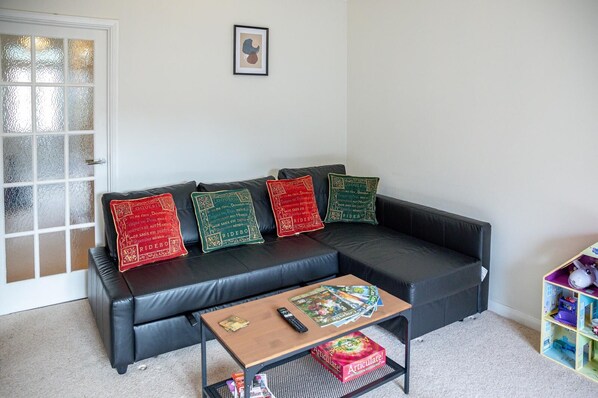 Una sala de estar con un sofá de cuero negro, tres almohadones decorativos, una mesa de centro con un juego de mesa y un tren de juguete.