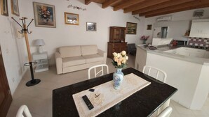 Een woonkamer met een bank, een salontafel met een bloevas, en een eethoek met stoelen. In de achtergrond is een keuken zichtbaar.