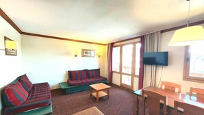 Ein Zimmer mit einem grünen Sofa, einem rot-schwarz gestreiften Sofa, einem Holztisch, einem Holzesstisch mit Stühlen, einem flachbildfernseher an der Wand und einem Fenster mit Vorhängen.