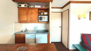 Eine kompakte Küche mit Holzschränken, einer Mikrowelle und einem Kühlschrank. Ein Esstisch mit Stuhl und einer kleinen Bank steht in der Ecke.