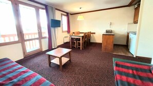 Ein Zimmer mit zwei Einzelbetten, einem kleinen Tisch, einem Schreibtisch und einem Stuhl. Es gibt ein Fenster mit Vorhängen, einen Fernseher und eine Lampe.