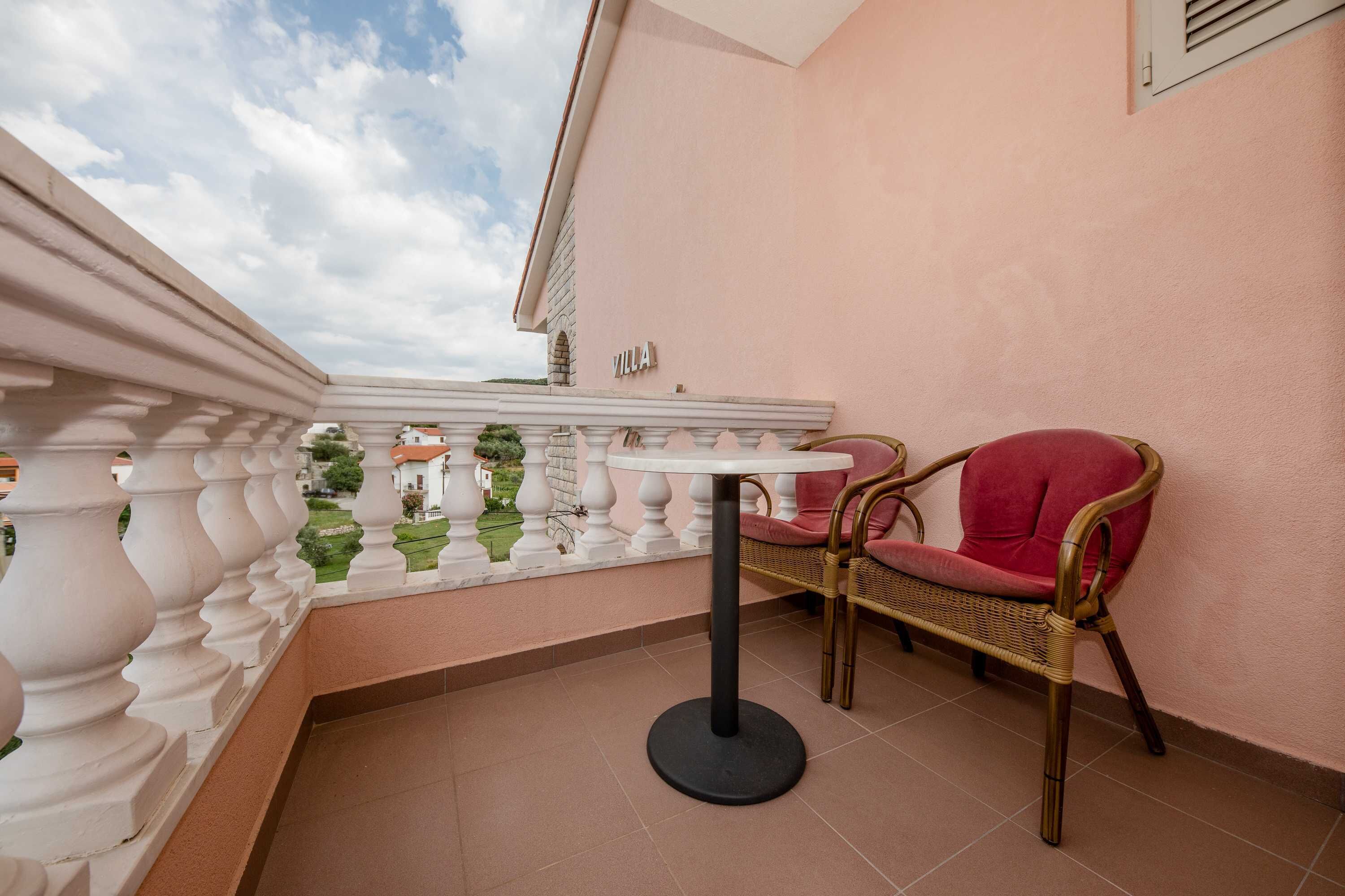 A balcony with a white balustrade, a wicker chair with a red cushion, and a round table.