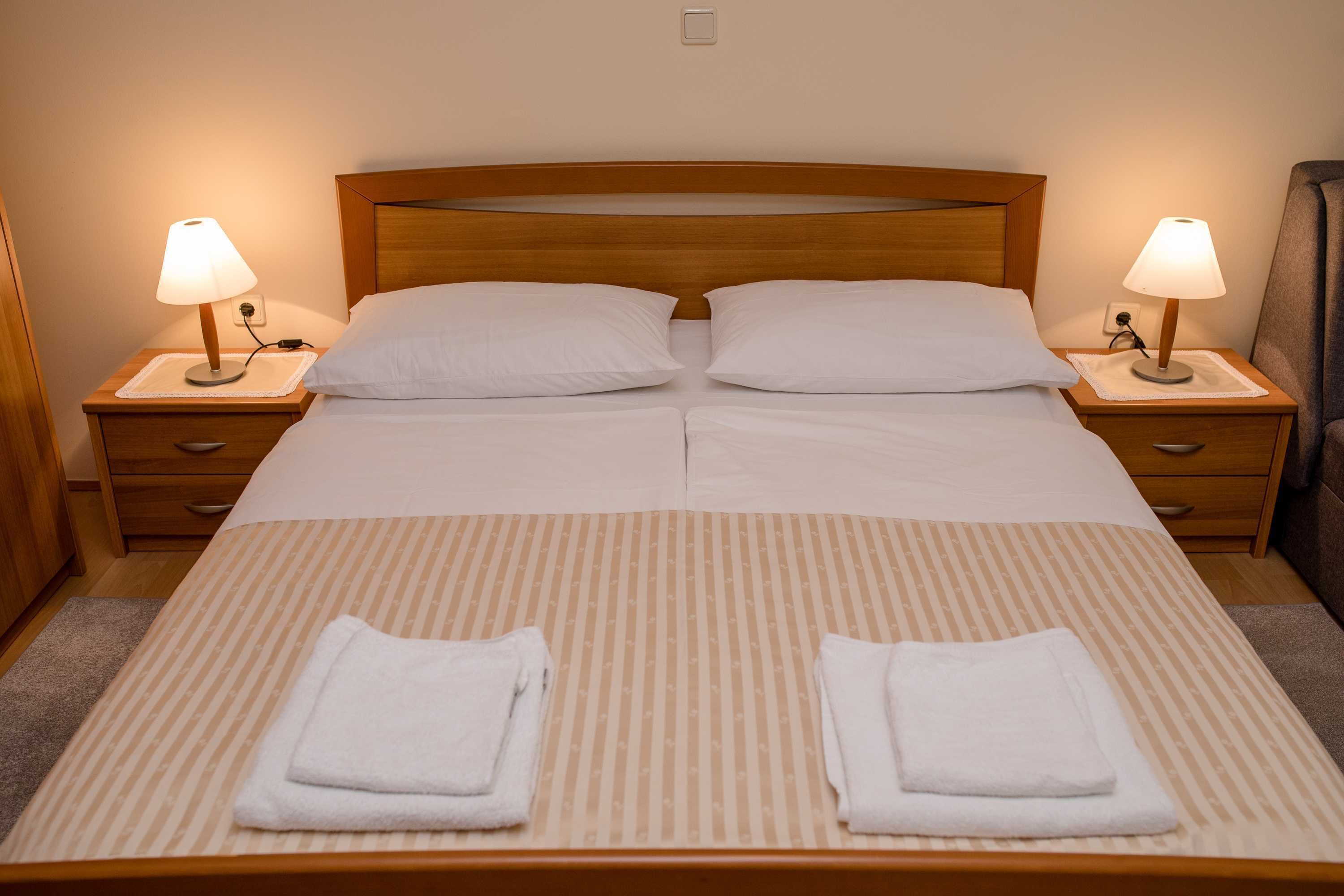 A double bed with two pillows, two bedside tables with lamps, and two folded towels on the bed.