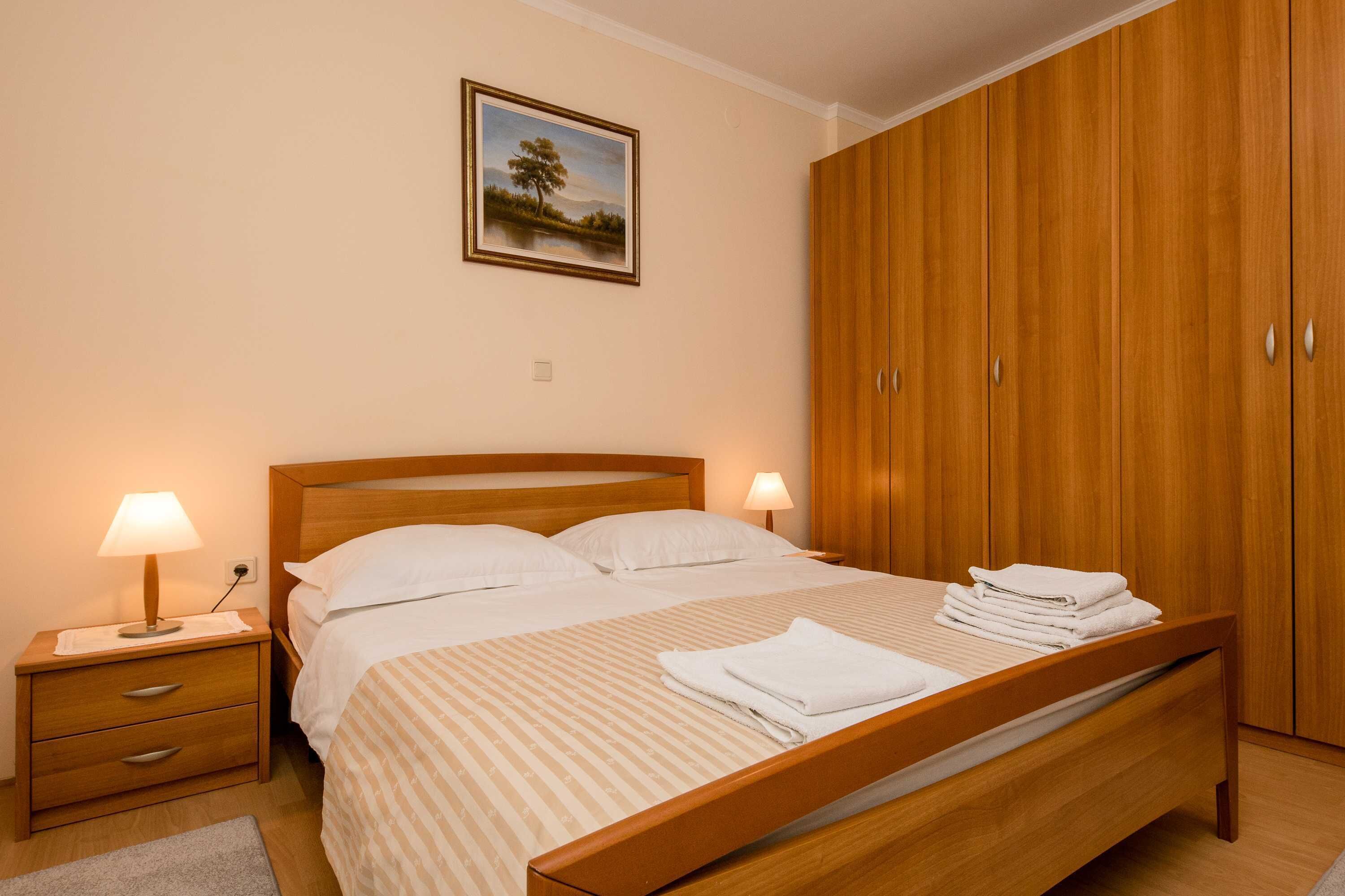 Un lit bien fait, avec du linge de lit blanc et une couverture à rayures, une table de chevet en bois avec une lampe, et une armoire avec un miroir.