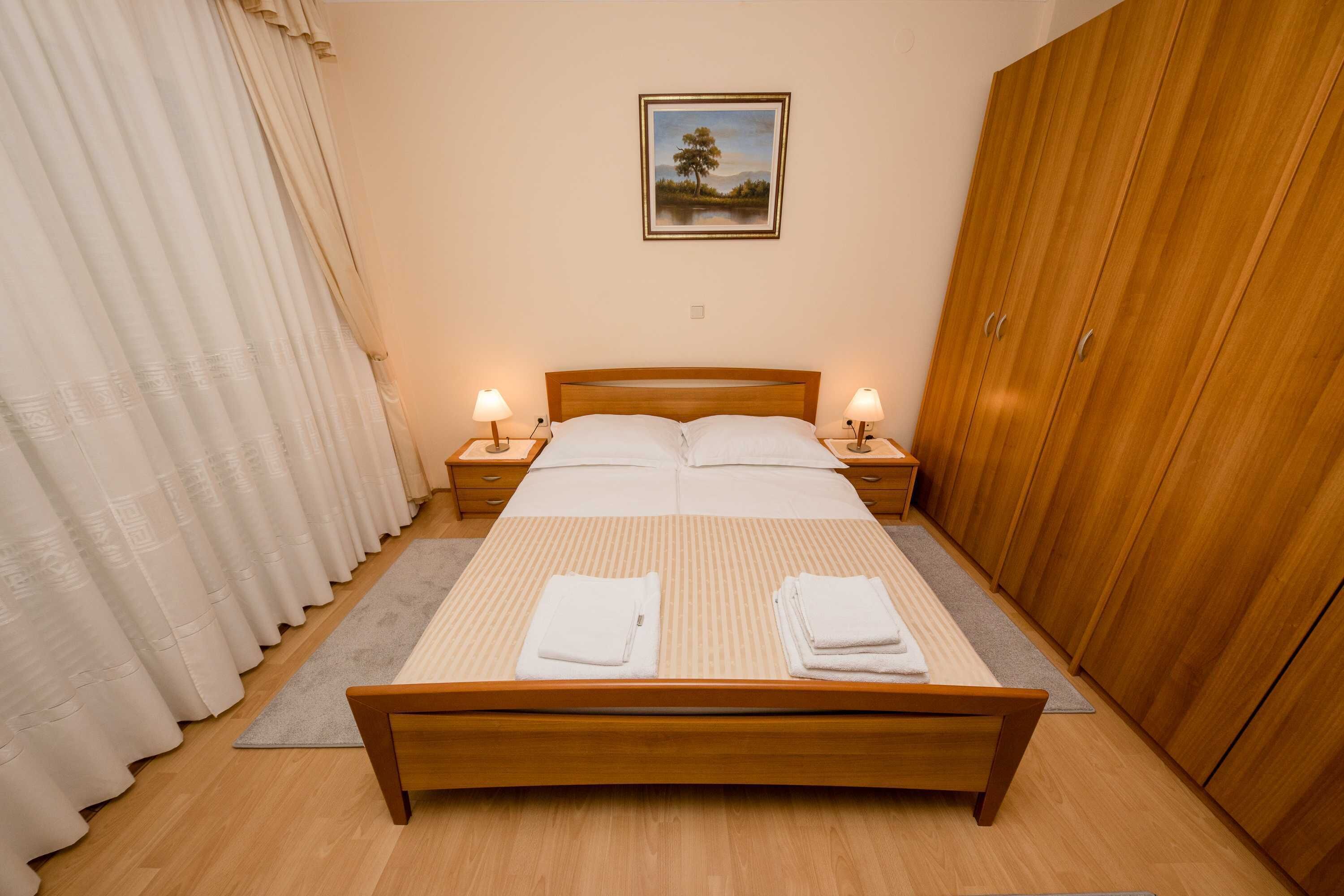 A neatly made bed with white linens and two pillows, flanked by bedside tables with lamps, in a room with wooden flooring and a large wardrobe.
