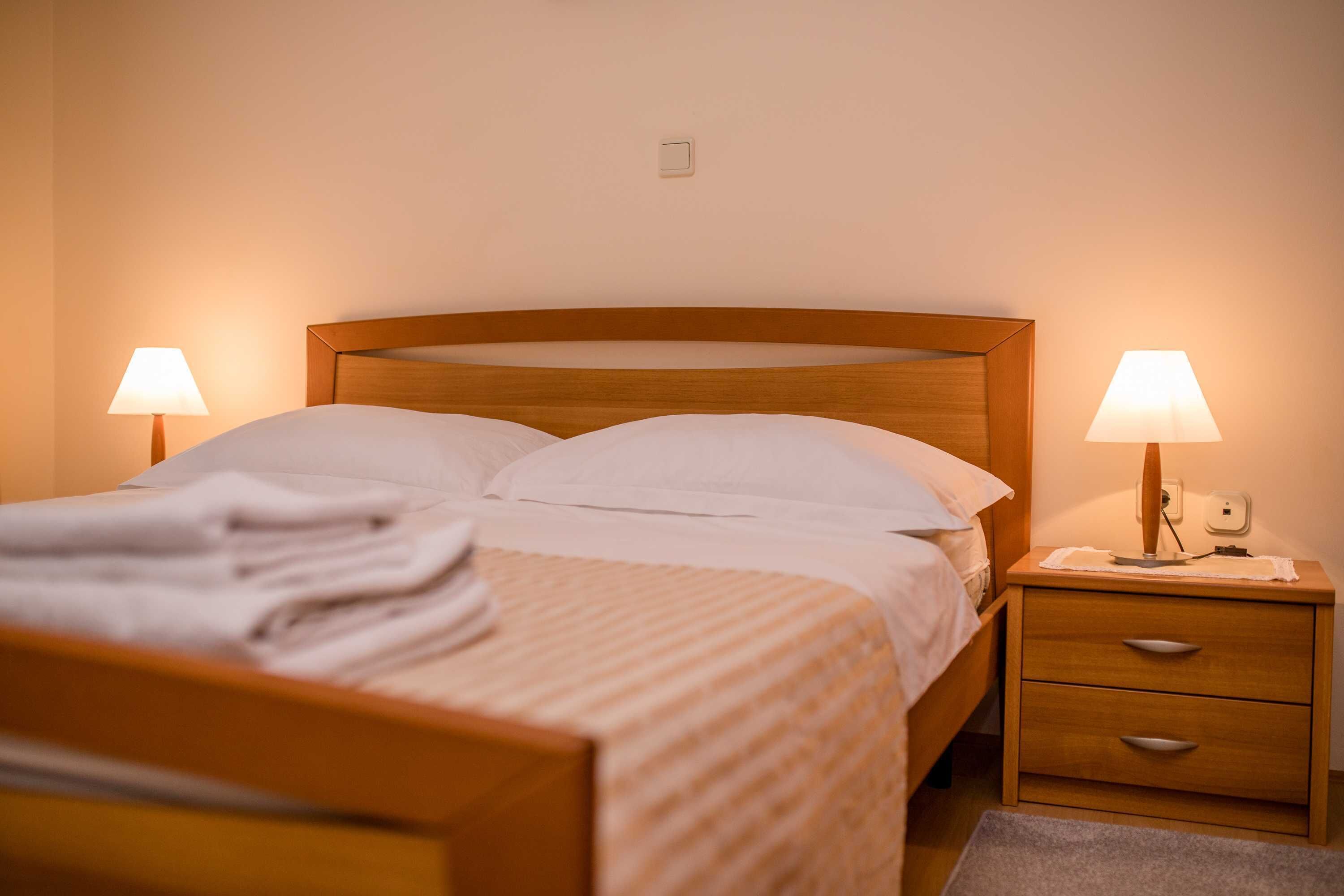 A neatly made bed with white linens and pillows, two bedside lamps, a wooden nightstand with drawers, and a small rug on the floor.