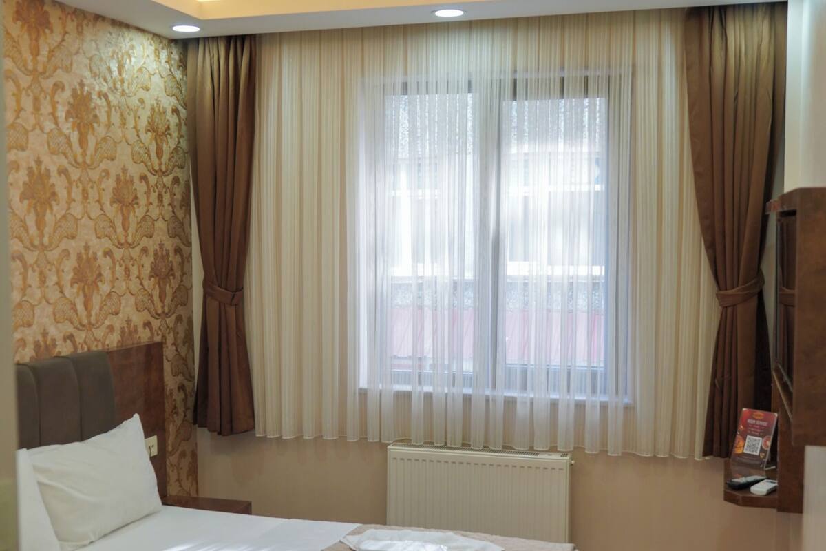A hotel room with a large bed, a radiator, a window with curtains, and a wall-mounted TV.