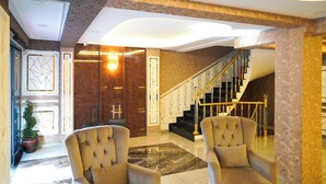 Un hall d’entrée spacieux avec un sol en marbre, deux fauteuils confortables, un escalier doté de rampes dorées et un grand lustre.
