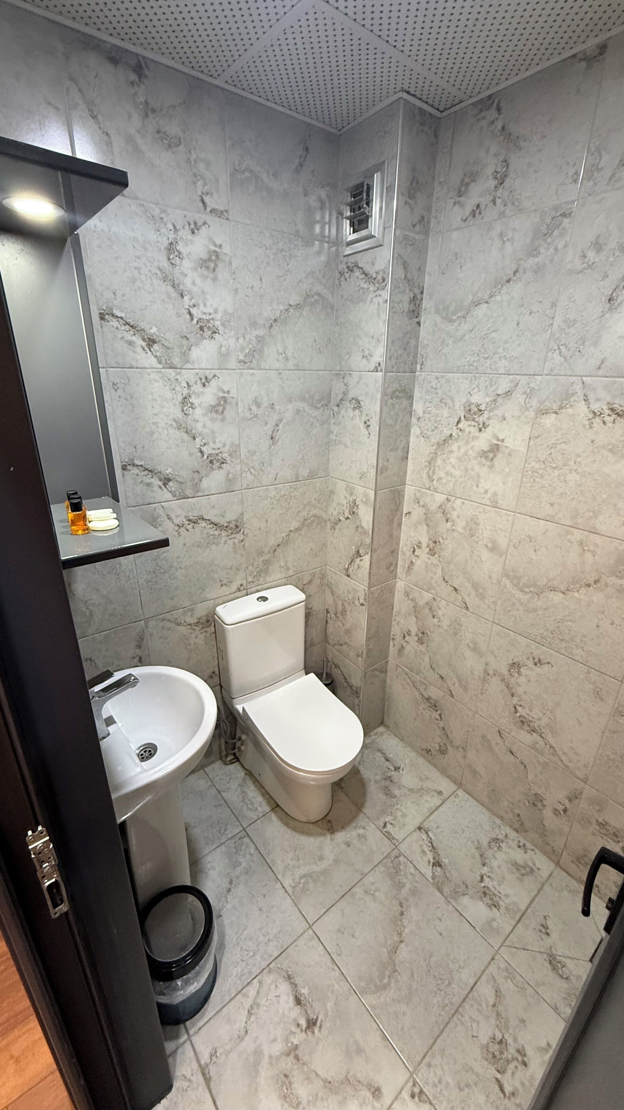 A bathroom with a toilet, sink, and shower area, featuring marble tiles and a ceiling with a perforated design.