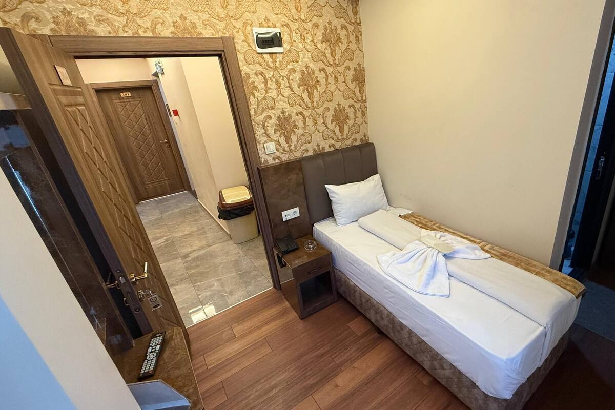 A hotel room with a bed, bedside table, and a small nightstand. There is a door leading to another room and a wall-mounted light fixture.