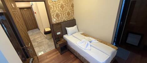 A hotel room with a bed, bedside table, and a small nightstand. There is a door leading to another room and a wall-mounted light fixture.