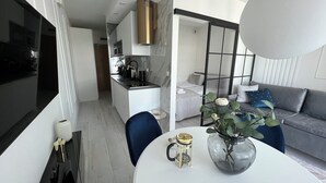 A modern kitchen with white cabinets, a black countertop, and a gold range hood. There is a dining area with a white table, a blue chair, and a vase with flowers. A gray sofa is visible in the background. - Apartament Glamour z basenem - 365PAM (Kolobrzeg)