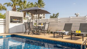 A swimming pool with a tiled edge, outdoor seating with a table and chairs, a grill, and palm trees in the background. - Protaras Villa Cemelia (Paralimni)