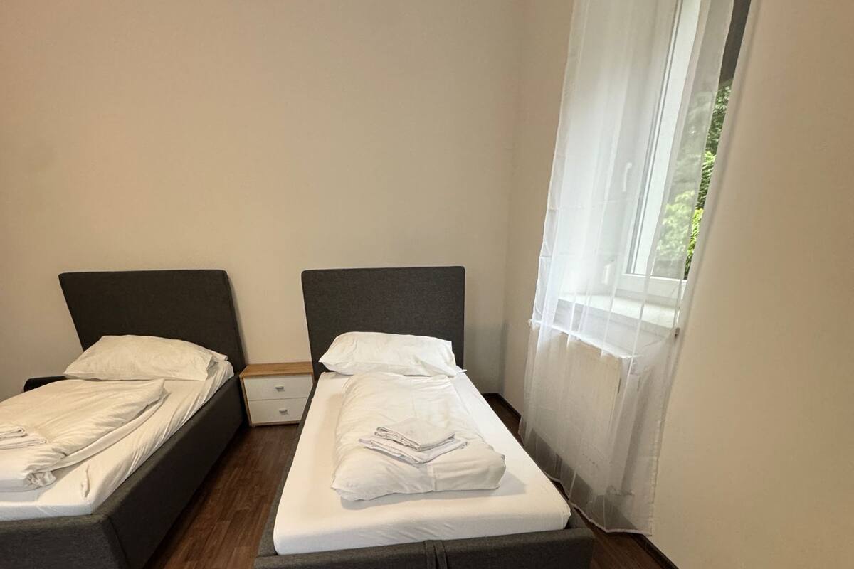 A room with two single beds, wooden flooring, and a window with sheer curtains.