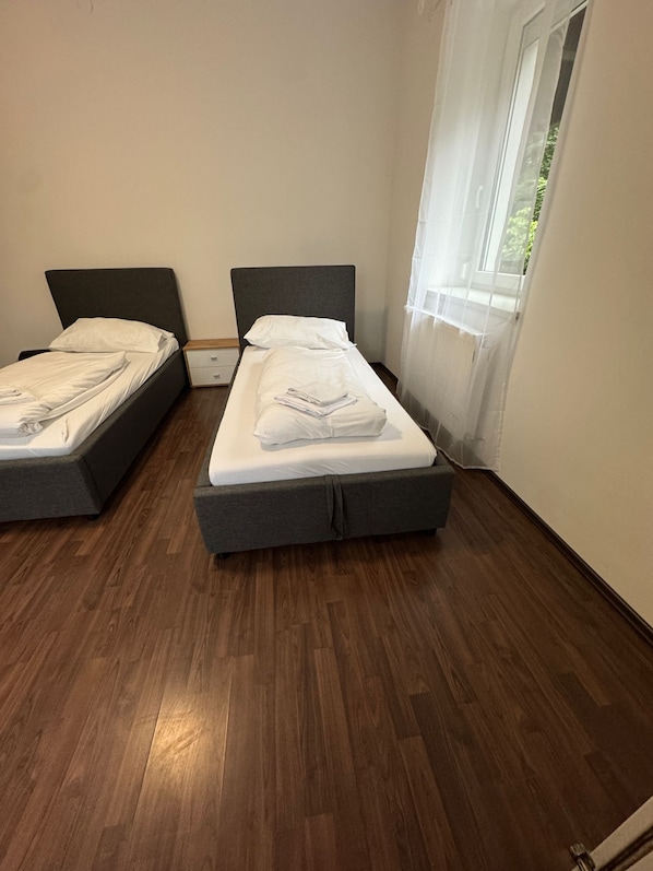 A room with two single beds, wooden flooring, and a window with sheer curtains.