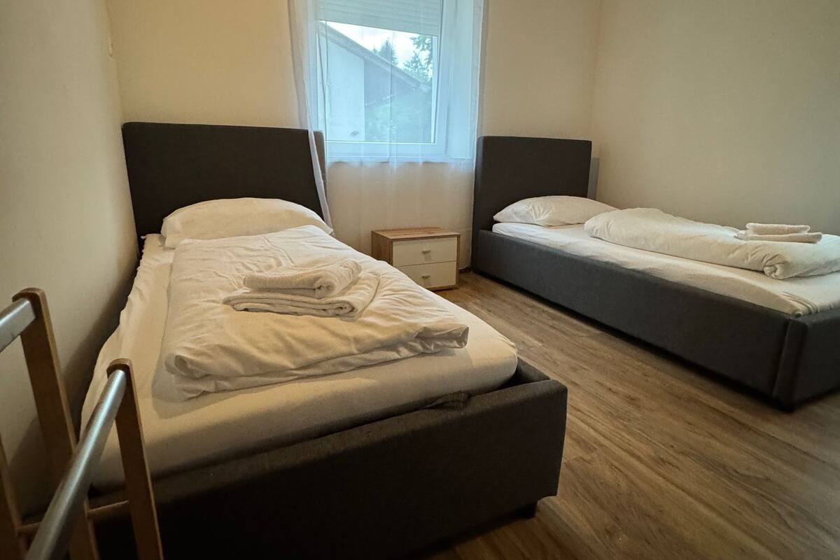 A room with two single beds, a wooden floor, a window with a view, and a small bedside table.