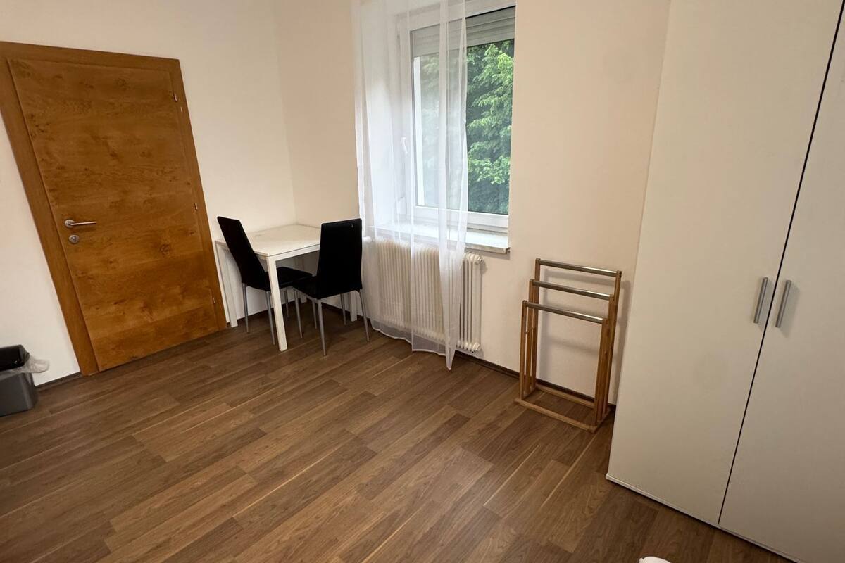 A room with a wooden floor, a bed, a desk with a chair, a radiator, and a wardrobe.