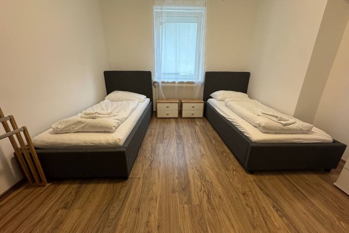 A small, well-lit room with two single beds, a wooden floor, a radiator, and a window with sheer curtains.