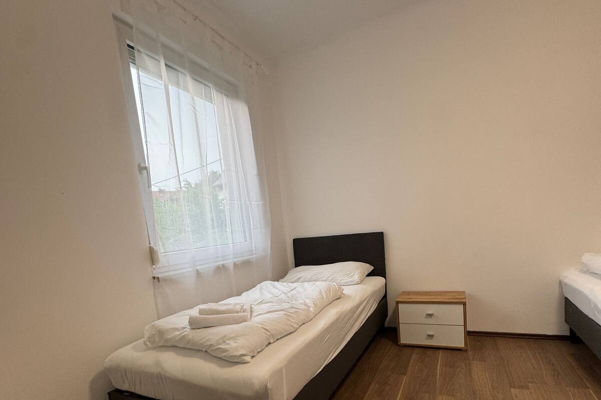 A bedroom with a single bed, a nightstand, and a window with sheer curtains.