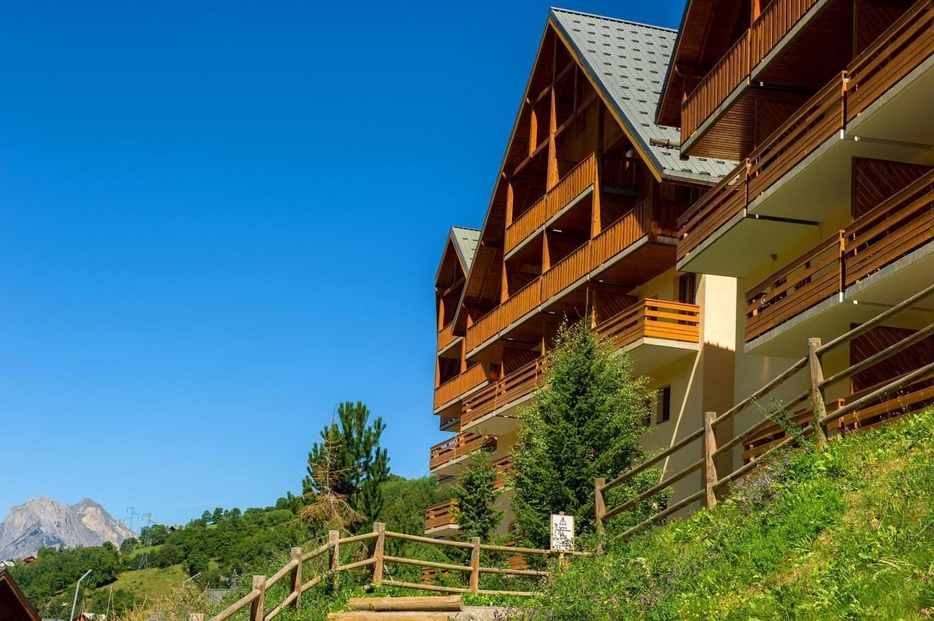 A multi-story wooden building with balconies, surrounded by greenery and a mountain in the background.