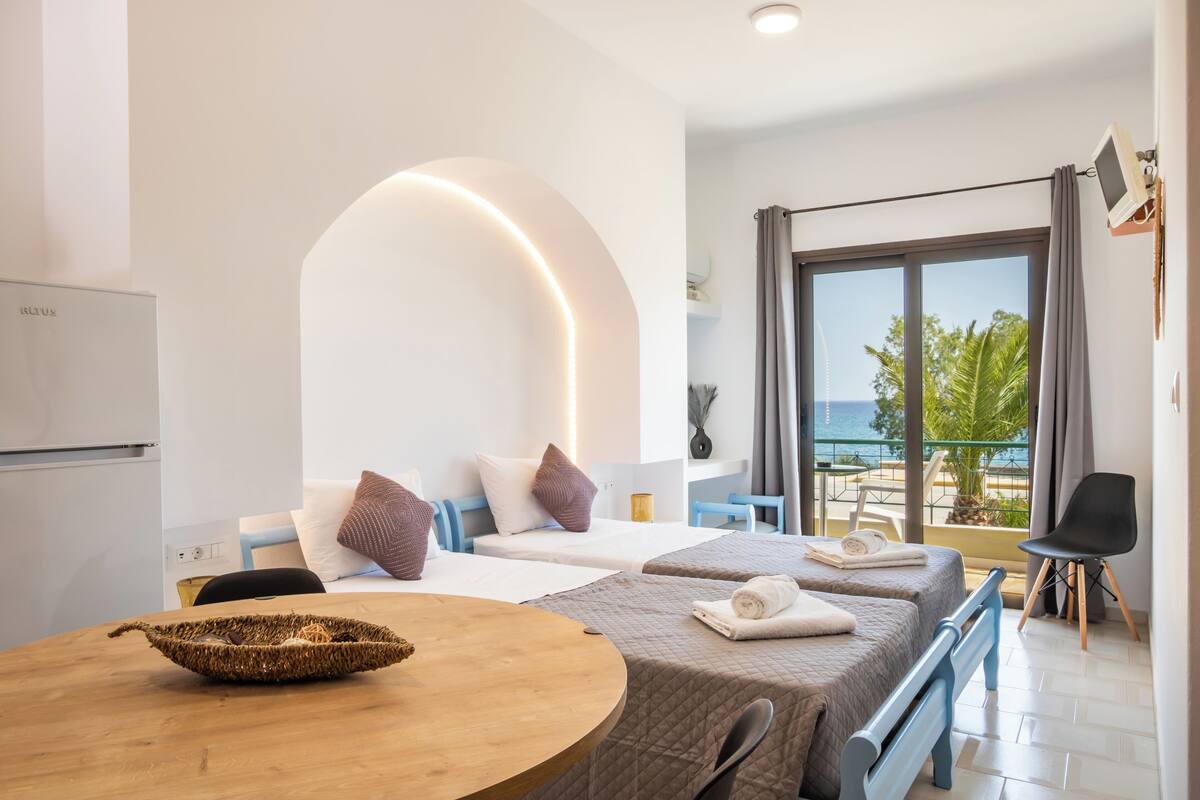 A hotel room with two beds, a balcony with a view of the sea, a dining table, and a TV mounted on the wall.