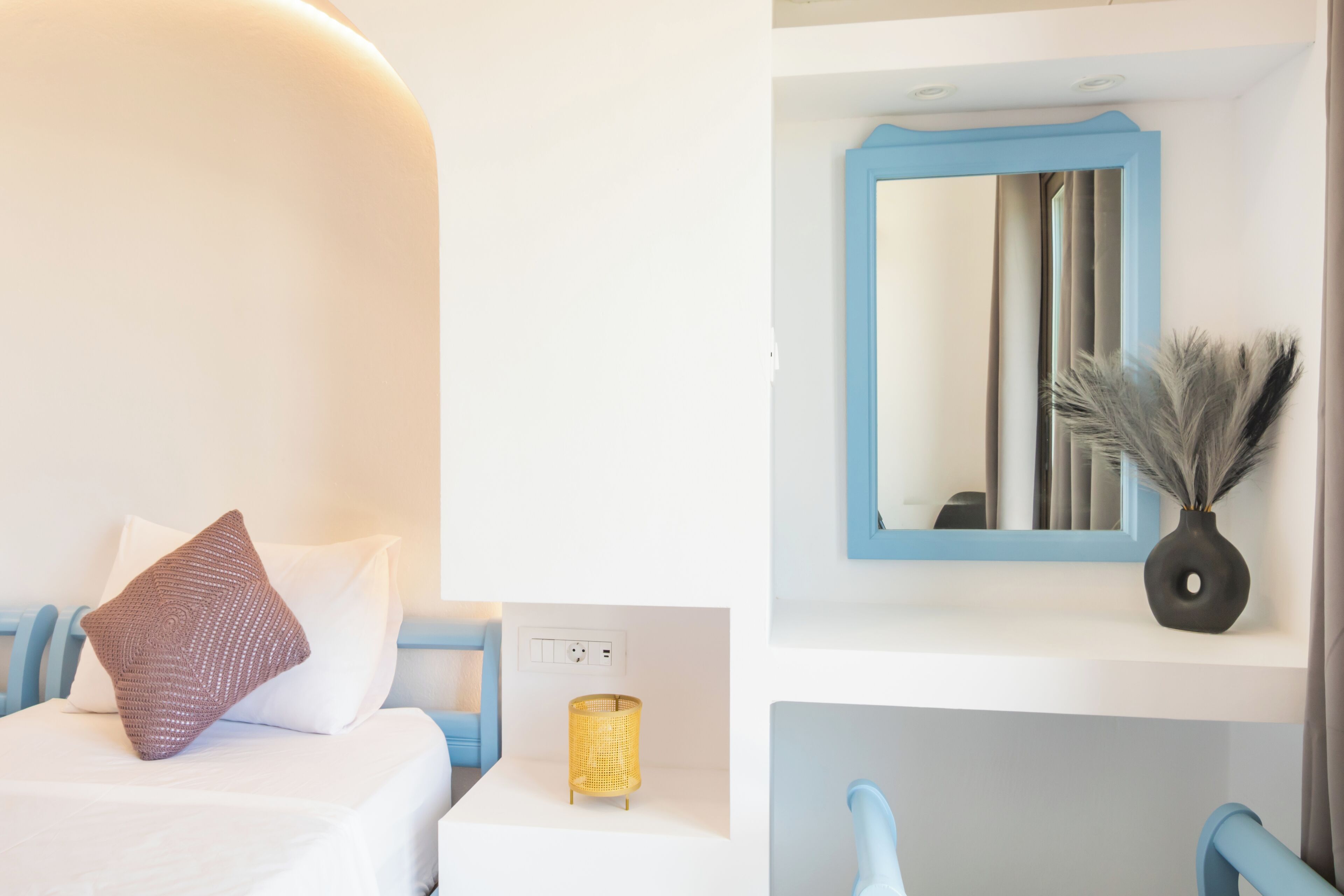 Une chambre d’hôtel avec un lit, un miroir, un vase décoratif contenant des branches et une prise électrique au mur.