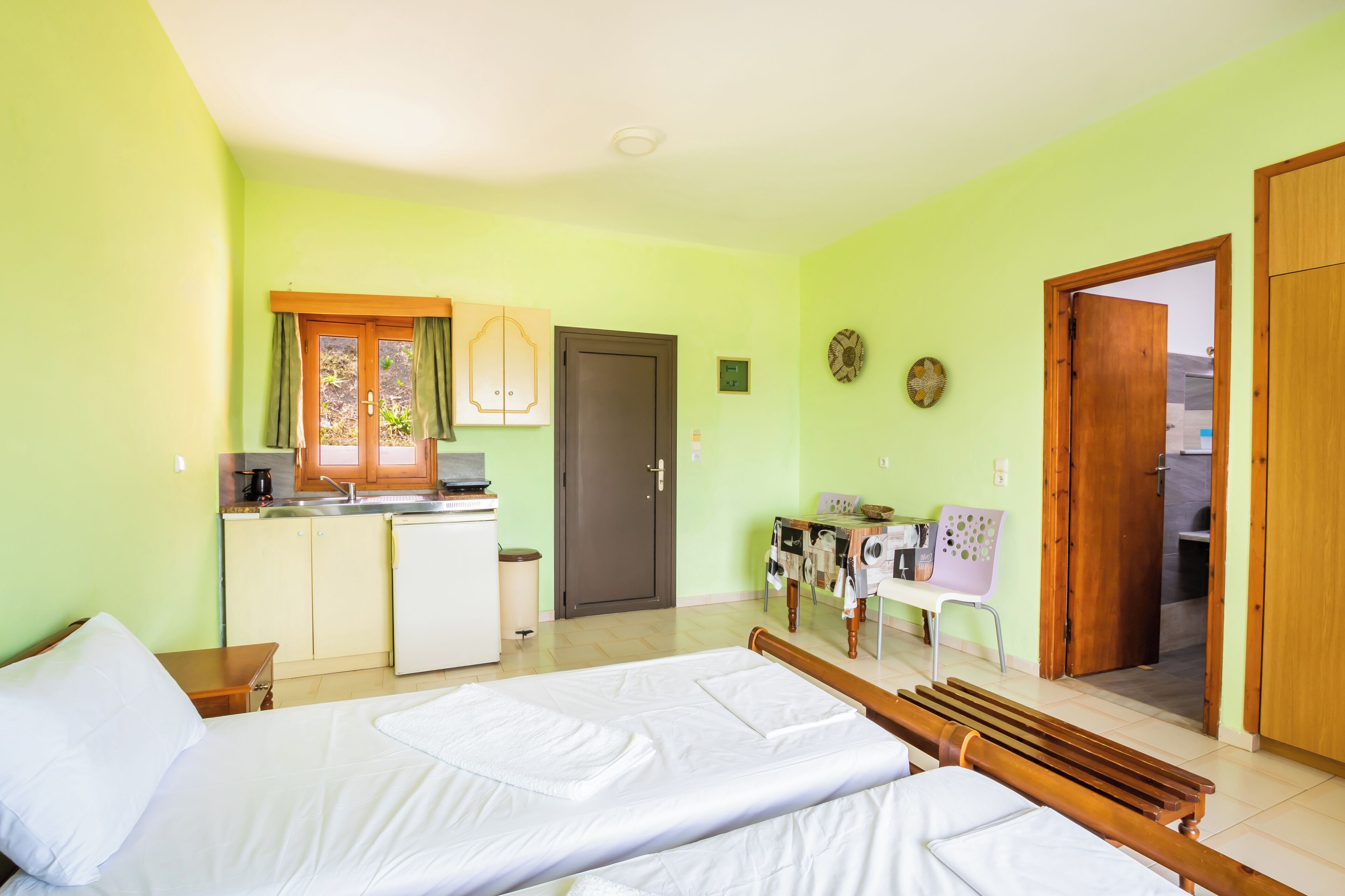 A room with two beds, a kitchenette, a desk, and a door leading to another room.
