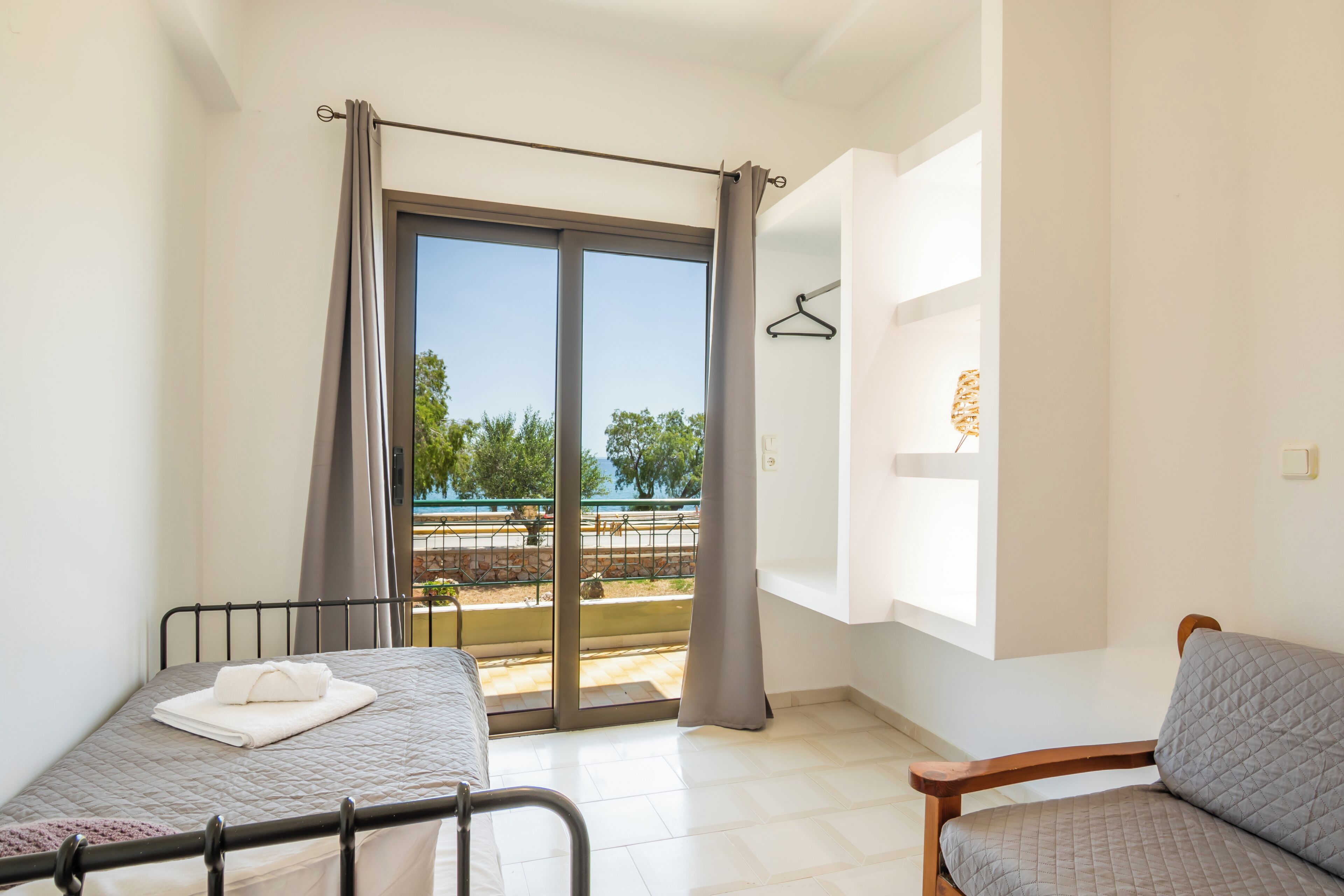 A room with a bed, a chair, a window with a view of a park, and a balcony.