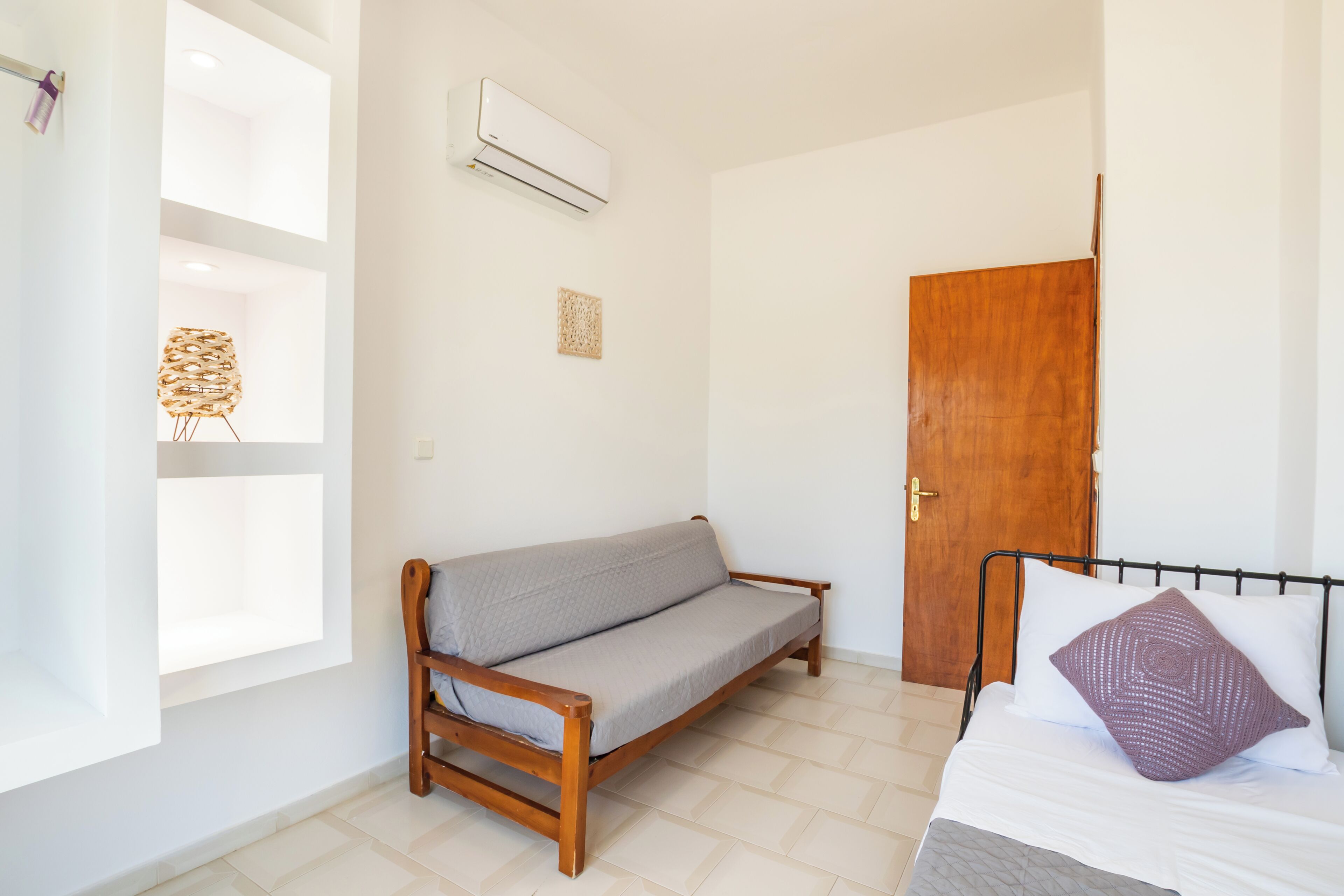 A room with a wooden sofa, a bed with a patterned pillow, a wooden door, and an air conditioning unit.