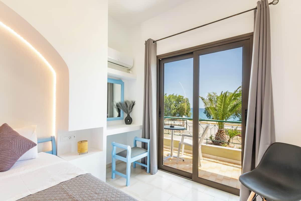 A bedroom with a bed, a desk, a chair, a balcony with a table and chairs, and a view of the sea.