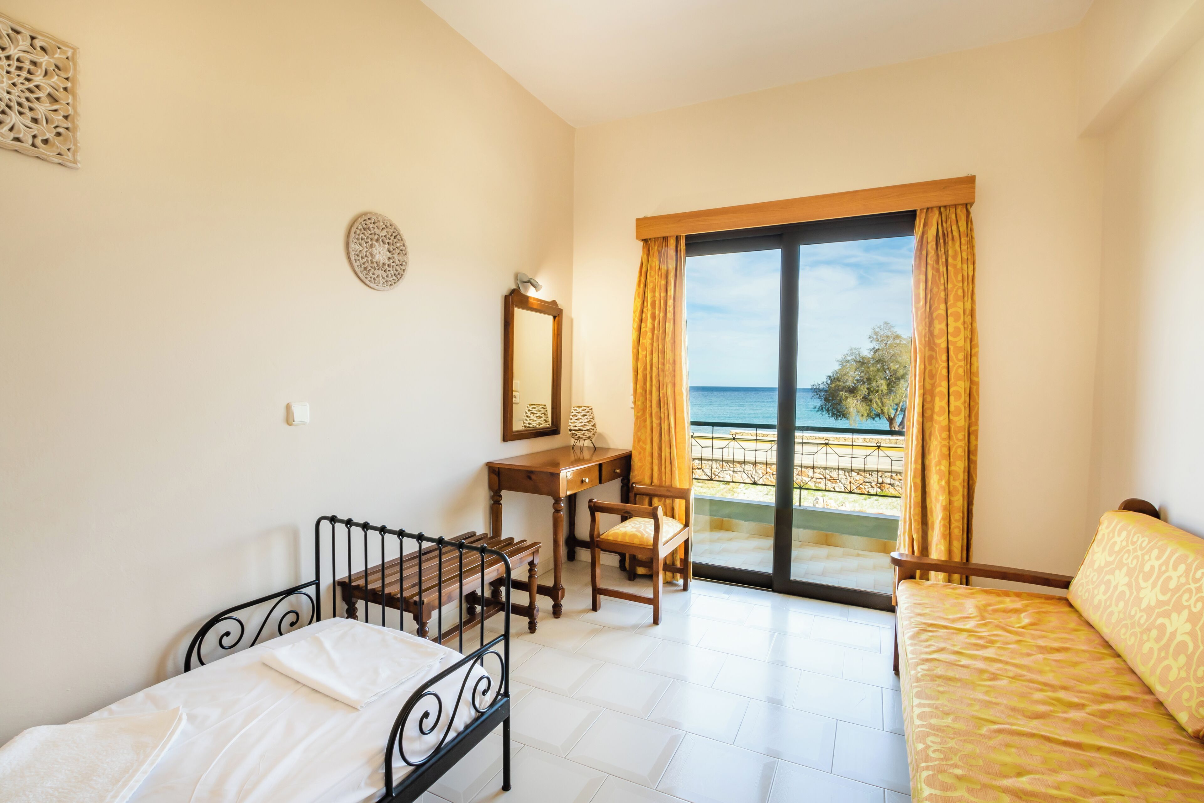 A bedroom with a bed, a desk, a chair, and a large window with a view of the sea.