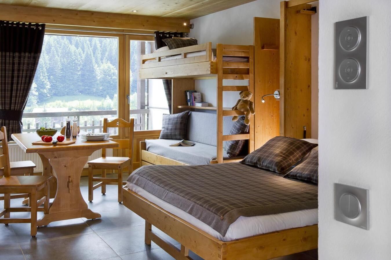 A cozy cabin-style room with a bunk bed, a wooden desk with a chair, and a window overlooking a forested area.