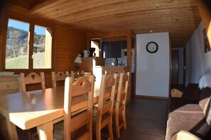 Un interior de madera con una mesa y sillas de comedor, una zona de cocina con un reloj, y vistas al exterior.