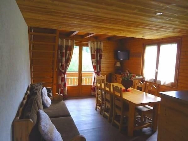 A wooden cabin interior with a dining table, chairs, a sofa, and a television.
