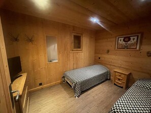 A wooden cabin room with a bed, a small wooden dresser, a television, and a window.