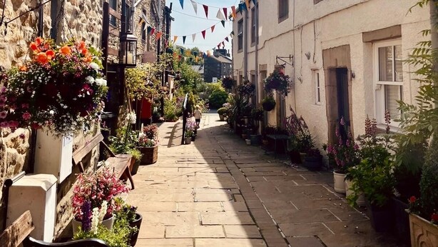 A narrow street lined with stone buildings, some with flower planters and hanging baskets. - The Old Middle School 1700 Cottage Addingham (Addingham)