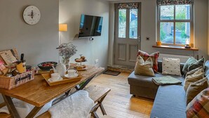 A cozy living room with a wooden dining table set for breakfast, a comfortable sofa with cushions, a television mounted on the wall, and a window with a view of greenery. - The Old Middle School 1700 Cottage Addingham (Addingham)