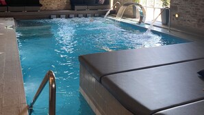 An indoor swimming pool with a stone wall backdrop, a lounge area with a bench, and a window with a view of greenery. - The Old Middle School 1700 Cottage Addingham (Addingham)